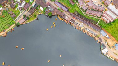 Göl kenarındaki bir köyün havadan görünüşü, kırsal mimari, yemyeşil ve sakin sulara dağılmış teknelerin karışımı sergileniyor. Doğal güzellik ve insan yerleşiminin ahenkli bir karışımı.