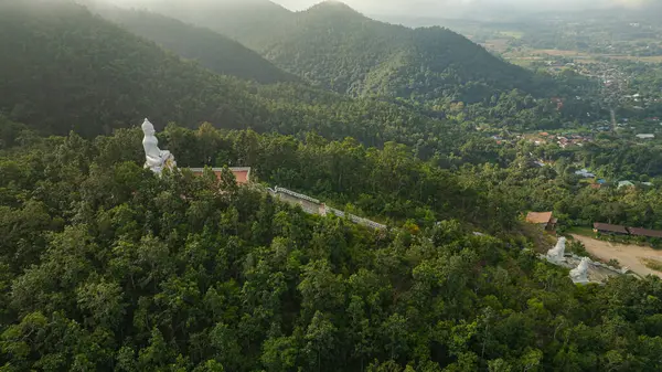Bereketli yeşilliklerle çevrili ve uzak köyleri olan huzurlu bir vadiye bakan ormanlık bir tepeye tünemiş büyük beyaz Buda heykelinin huzurlu bir hava görüntüsü. Huzurlu ortam ruhani ve doğal uyum yayar..  