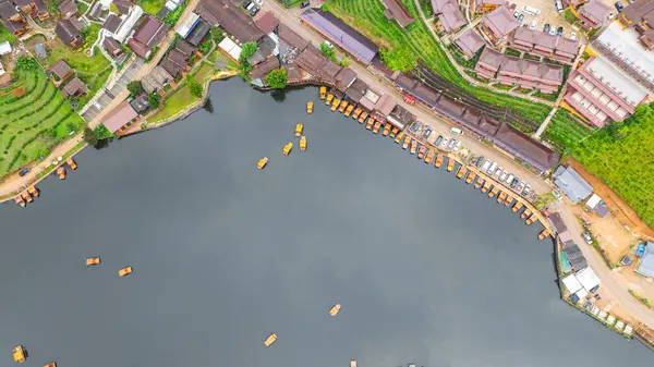 Göl kenarındaki bir köyün havadan görünüşü, kırsal mimari, yemyeşil ve sakin sulara dağılmış teknelerin karışımı sergileniyor. Doğal güzellik ve insan yerleşiminin ahenkli bir karışımı.