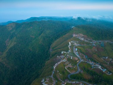 Bereketli yeşillikler, dağınık evler ve tarım arazileriyle çevrili dolambaçlı bir dağ yolunun havadan görünüşü. Bu manzaralı manzara, dağlık bir bölgedeki doğal güzellikleri ve insan yerleşimlerini yansıtıyor..  