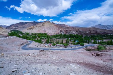 Aşağıdaki vadide yemyeşil ağaçlarla dolu kurak kayalık bir araziyi kesen dolambaçlı bir dağ yolunun manzarası. Dağınık bulutlar ve uzak karlarla dolu dramatik bir mavi gökyüzüne karşı kurulmuş.