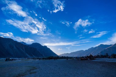 Dramatik bir dağ vadisinde nefes kesici bir gün batımı. Huzurlu manzara renkli gökyüzü ile zenginleşir. Uzak deve sürüleri ve yükselen zirveler doğayla bir barış ve bağ hissi uyandırıyor..