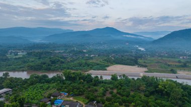 Şafak vakti geniş bir kırsal alanda akan dolambaçlı bir nehrin çarpıcı hava manzarası. Yumuşak sabah ışığı ve sisli atmosfer dağınık köylerle sakin ve resim gibi bir manzara yaratır.