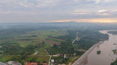 Şafak vakti geniş bir kırsal alanda akan dolambaçlı bir nehrin çarpıcı hava manzarası. Yumuşak sabah ışığı ve sisli atmosfer dağınık köylerle sakin ve resim gibi bir manzara yaratır.