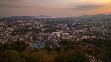 Gün batımında yemyeşil dağların arasına yerleşmiş bir kasabanın manzarası. Tepenin yamacına tünemiş bir tapınak yapısı ve canlı turuncu bir gökyüzü ılık, sakin bir atmosfer yaratıyor..