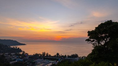 Phuket, Tayland 'daki Kata Sahili' nin gün batımında çekilen nefes kesici hava görüntüsü. Görüntü kıyı şeridini, küçük adaları, yemyeşil tepeleri ve parlak bir gökyüzünün altındaki canlı şehri gösteriyor.