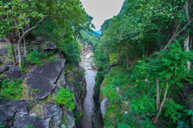 Yükselen kaya oluşumları ve uçurumları birbirine bağlayan dar bir köprüyle, yemyeşil ormanlarla çevrili bir nehir vadisinin pitoresk görüntüsü. Doğal güzellik ve canlı yeşillik. Öpücük uçurumu