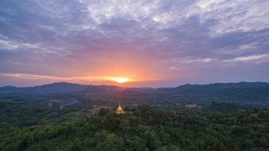 Parlak bir günbatımında yemyeşil tepelerin arasında yuva yapmış, çarpıcı bir hava manzarası. Dramatik bulutlar ve yumuşak güneş ışığı manzaranın üzerine huzur dolu bir parıltı saçıyor..