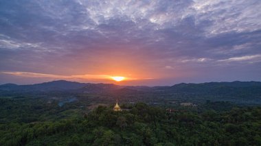 Parlak bir günbatımında yemyeşil tepelerin arasında yuva yapmış, çarpıcı bir hava manzarası. Dramatik bulutlar ve yumuşak güneş ışığı manzaranın üzerine huzur dolu bir parıltı saçıyor..