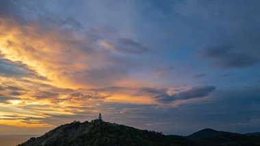 Nefes kesen bir günbatımı, kıyı kasabasının üzerinde turuncu ve mavi renkli canlı renkler çiziyor. Sahneyi çevreleyen yemyeşil tepeler var. Uzak bir tepe yapısı huzur dolu bir manzara ekler..  