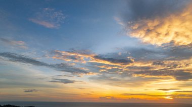 Nefes kesen bir günbatımı, kıyı kasabasının üzerinde turuncu ve mavi renkli canlı renkler çiziyor. Sahneyi çevreleyen yemyeşil tepeler var. Uzak bir tepe yapısı huzur dolu bir manzara ekler..  