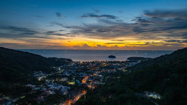 Nefes kesen bir günbatımı, kıyı kasabasının üzerinde turuncu ve mavi renkli canlı renkler çiziyor. Sahneyi çevreleyen yemyeşil tepeler var. Uzak bir tepe yapısı huzur dolu bir manzara ekler..  