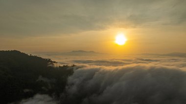 Yoğun bulutlarla kaplı bir dağ vadisinde yükselen güneşin nefes kesici hava manzarası. Altın ışık sisi aydınlatıyor, şafakta dramatik ve ruhani bir atmosfer yaratıyor.