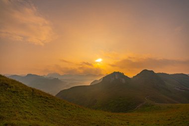 Sakin günbatımının havadan görünüşü, yuvarlanan tepeler ve uzak dağ tepeleri üzerinde altın bir parıltı oluşturarak, sisli ve yumuşak ışık katmanlarından oluşan barışçıl ve atmosferik bir manzara yaratır. dağ arkaplanı.