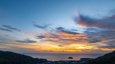 Nefes kesen bir günbatımı, kıyı kasabasının üzerinde turuncu ve mavi renkli canlı renkler çiziyor. Sahneyi çevreleyen yemyeşil tepeler var. Uzak bir tepe yapısı huzur dolu bir manzara ekler..  