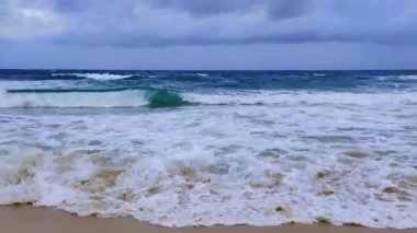 Dalgalar bulutlu gökyüzünün altında kumlu sahile çarpıyor.