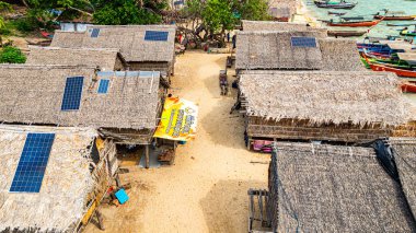 Phang Nga, Tayland-Mart, 20.2025: Yeşillik yeşil ormanlarla çevrili kıyı şeridi boyunca sıralanmış küçük kulübeleriyle tropikal bir sahil Morgan köyünün çarpıcı hava manzarası. Balıkçı tekneleri demirli.