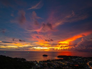 Nefes kesen bir günbatımı, kıyı kasabasının üzerinde turuncu ve mavi renkli canlı renkler çiziyor. Sahneyi çevreleyen yemyeşil tepeler var. Uzak bir tepe yapısı huzur dolu bir manzara ekler..  