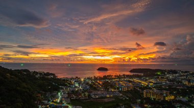 Nefes kesen bir günbatımı, kıyı kasabasının üzerinde turuncu ve mavi renkli canlı renkler çiziyor. Sahneyi çevreleyen yemyeşil tepeler var. Uzak bir tepe yapısı huzur dolu bir manzara ekler..  