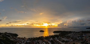 Nefes kesen bir günbatımı, kıyı kasabasının üzerinde turuncu ve mavi renkli canlı renkler çiziyor. Sahneyi çevreleyen yemyeşil tepeler var. Uzak bir tepe yapısı huzur dolu bir manzara ekler..  