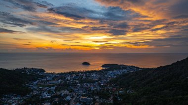 Nefes kesen bir günbatımı, kıyı kasabasının üzerinde turuncu ve mavi renkli canlı renkler çiziyor. Sahneyi çevreleyen yemyeşil tepeler var. Uzak bir tepe yapısı huzur dolu bir manzara ekler..  