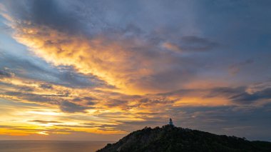 Nefes kesen bir günbatımı, kıyı kasabasının üzerinde turuncu ve mavi renkli canlı renkler çiziyor. Sahneyi çevreleyen yemyeşil tepeler var. Uzak bir tepe yapısı huzur dolu bir manzara ekler..  