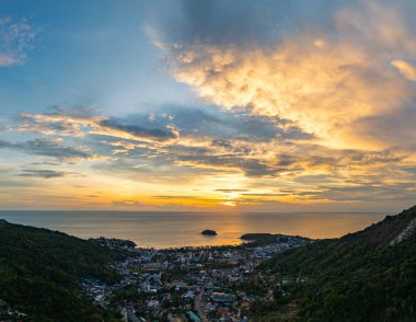 Nefes kesen bir günbatımı, kıyı kasabasının üzerinde turuncu ve mavi renkli canlı renkler çiziyor. Sahneyi çevreleyen yemyeşil tepeler var. Uzak bir tepe yapısı huzur dolu bir manzara ekler..  