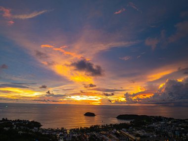 Nefes kesen bir günbatımı, kıyı kasabasının üzerinde turuncu ve mavi renkli canlı renkler çiziyor. Sahneyi çevreleyen yemyeşil tepeler var. Uzak bir tepe yapısı huzur dolu bir manzara ekler..  