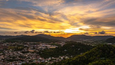 Gökyüzünü, Khao Rang 'ın üzerindeki altın ve turuncu renklerle dolduran, tepelerin doğal hatlarını ve aşağıdaki kentsel manzaranın sessiz çekiciliğini vurgulayan, ışıl ışıl bir gün batımı manzarası.