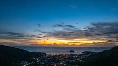 Nefes kesen bir günbatımı, kıyı kasabasının üzerinde turuncu ve mavi renkli canlı renkler çiziyor. Sahneyi çevreleyen yemyeşil tepeler var. Uzak bir tepe yapısı huzur dolu bir manzara ekler..  