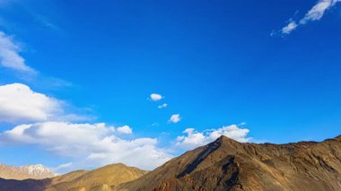 Yumuşak beyaz bulutlarla dağılmış canlı mavi gökyüzünün altındaki engebeli dağların nefes kesici manzarası. Görüntü, yüksek rakımlı bir arazinin uçsuz bucaksızlığını ve huzurunu yakalar..