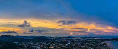 Nefes kesen bir gün batımının hava hiper yanılma görüntüsü sahneyi çevreleyen yemyeşil tepelerle dolu kıyı kasabasının üzerinde turuncu ve mavi renkli gökyüzünü resmediyor. Okyanusun üzerinde dramatik gün batımı bulutu