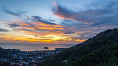 Nefes kesen bir günbatımı, kıyı kasabasının üzerinde turuncu ve mavi renkli canlı renkler çiziyor. Sahneyi çevreleyen yemyeşil tepeler var. Uzak bir tepe yapısı huzur dolu bir manzara ekler..  
