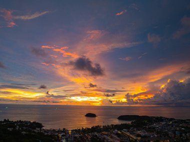 Nefes kesen bir günbatımı, kıyı kasabasının üzerinde turuncu ve mavi renkli canlı renkler çiziyor. Sahneyi çevreleyen yemyeşil tepeler var. Uzak bir tepe yapısı huzur dolu bir manzara ekler..  