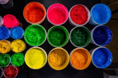 A collection of plastisol inks stored in sturdy barrels and glass jars, each filled with rich and vibrant colors red, blue, yellow, green, pink, orange, purple, and white in screen printing factory