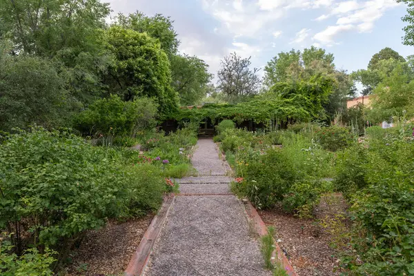 A gravel walking path lined with bushes and wildflowers leads to an ivy covered entryway in a tranquil garden, with green trees and a partly cloudy sky in the background.