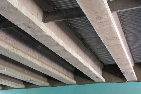 Architectural detail of concrete support beams beneath an overpass, with metal sheeting between the beams, and strong shadows.