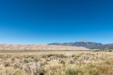 Açık mavi gökyüzünün altındaki çalı çayırları, kum tepeleri ve dağlardan oluşan çöl manzarası Colorado 'daki Great Sand Dunes Ulusal Parkı' nın dışında çekilmiştir..