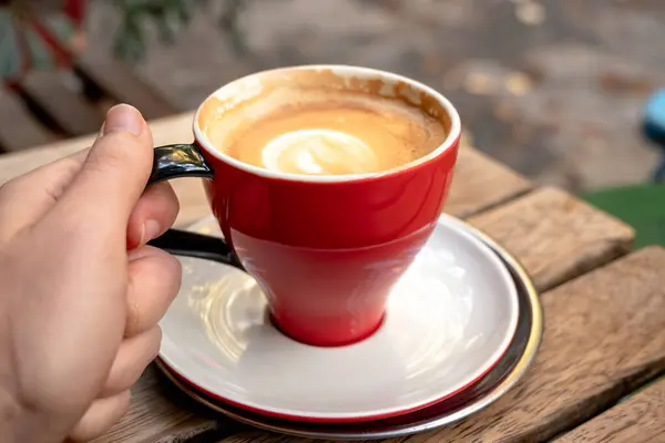  Bir adam kırmızı fincanı elinde latte kahveyle ahşap bir masada tutuyor..