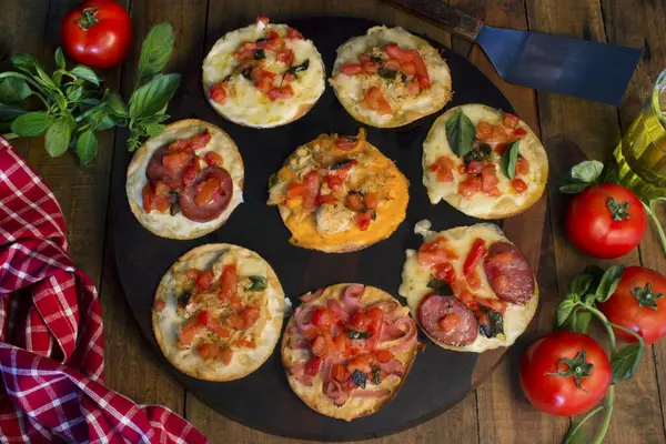 Small cheese and pepperoni pizzas with tomatoes, on a wooden board and table, in production a red and white checkered cloth, tomatoes and seasonings, a glass cup with olive oil and a spatula