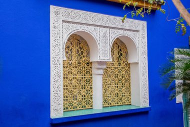 Canlı bir mavi duvar, süslü bir Fas tarzı pencereyi, karmaşık beyaz alçı kemerleri ve dekoratif altın demiri, egzotik işçiliği ve mimari güzelliği ile çerçeveler..