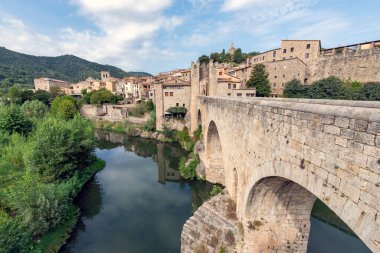 Bir nehrin üzerindeki köprü mavi gökyüzü ve bulutları olan bir ortaçağ şehrine erişim sağlıyor.