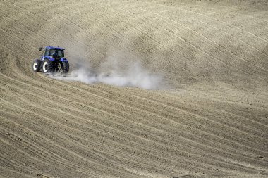 Bir traktör yeni sürülmüş dalgalı topraklarda hareket ederek, minimalist bir arazide tarım makinelerini ve kırsal verimliliği vurguluyor.