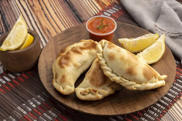 Homemade empanadas cooked with chicken meat vegetables and ham and cheese on a wooden plate with sauces and lemon