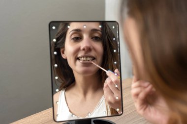 Güzellik ve makyaj merkezinde makyaj yapan genç bir kadın ayna ve cep telefonunun önünde mutlu bir yüzükle dudaklarını boyuyor ve yüz güzelliği için elmacık kemiklerini boyuyor. 