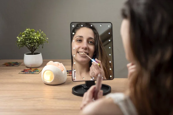 young woman in beauty and makeup center putting on makeup in front of a mirror and cell phone with a ring of light happy painting her lips and cheekbones for facial beauty