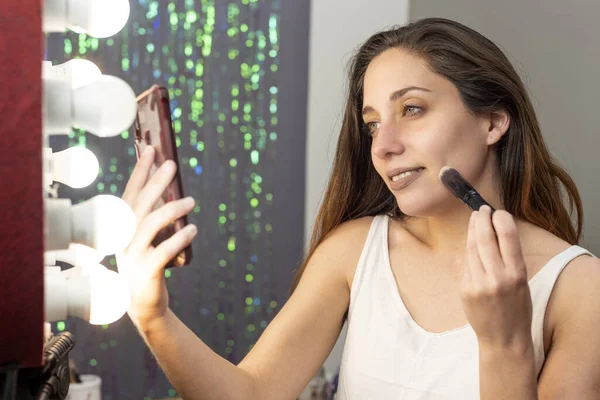 young woman in beauty and makeup center putting on makeup in front of a mirror and cell phone with a ring of light happy painting her lips and cheekbones for facial beauty