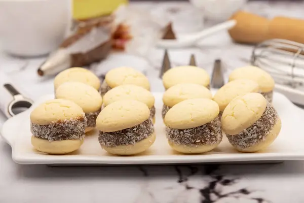 stock image Homemade cornstarch alfajores with dulce de leche and grated coconut on a kitchen counter with the ingredients flour sugar coconut whisk