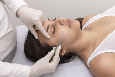 young woman on a table in a beauty center performing a beauty treatment for facial skin with a scalpel using the dermaplaning technique  clipart