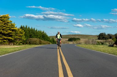 Orman ve dağların arka planına sahip bir yolun ortasında bisikletini süren bir erkek bisikletçi. 